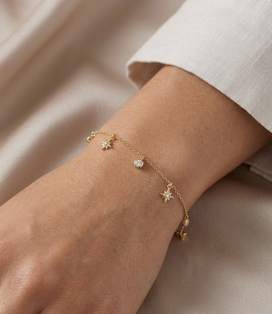 Gold bracelet with star and heart charms on a wrist against a neutral background